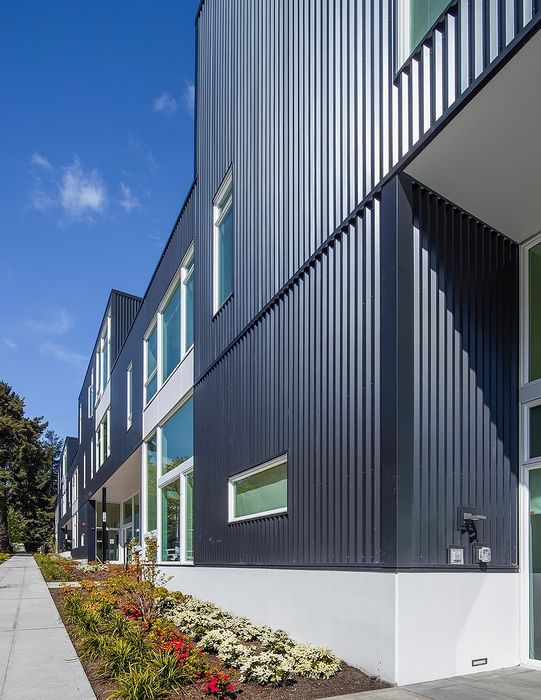 Modern apartment exterior featuring sleek black metal panels, large windows, and vibrant landscaped surroundings under a clear blue sky.