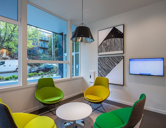 Modern apartment lounge with vibrant green and yellow chairs, large windows offering a lush view, and contemporary wall art.