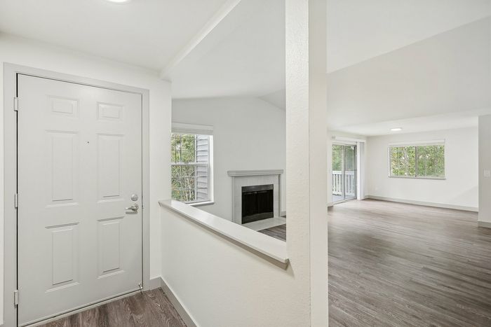 Bright and inviting open-concept interior with hardwood floors, large windows, and a cozy fireplace leading to a spacious balcony.