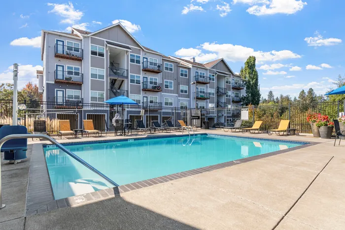 Inviting apartment complex with a sparkling pool, sunlit lounge chairs, and charming balconies against a clear blue sky.
