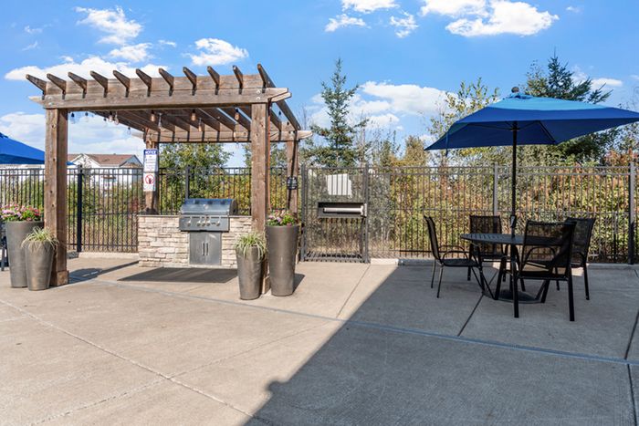 Inviting outdoor space featuring a wooden pergola with a built-in grill, surrounded by lush greenery and complemented by modern patio seating with blue umbrellas.