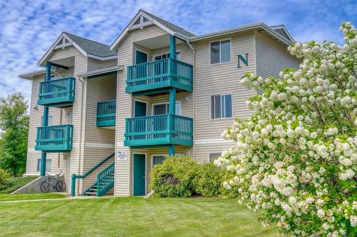Charming apartment exterior with bright teal balconies, situated amidst lush green lawns and blooming white flowers.