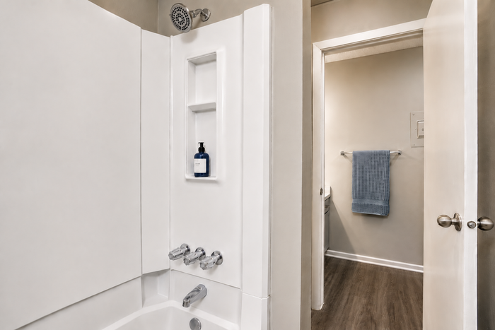 Elegant bathroom with modern bathtub, sleek fixtures, and convenient built-in shelving, leading to a warmly lit washroom with wooden flooring.