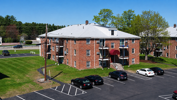 Charming brick apartment building with ample parking, lush green surroundings, and an inviting entrance canopy.