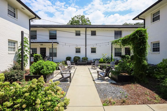Charming courtyard with inviting seating, lush greenery, and string lights between modern white brick buildings under a clear blue sky.