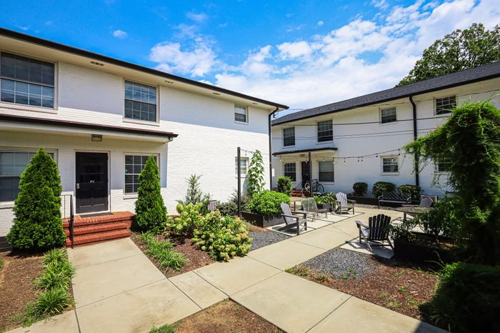 Charming apartment complex courtyard with modern white brick buildings, inviting outdoor seating area, and lush green landscaping under a bright blue sky.