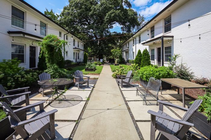 Charming outdoor courtyard with lush greenery, cozy seating, and elegant string lighting between two stylish white brick apartment buildings.
