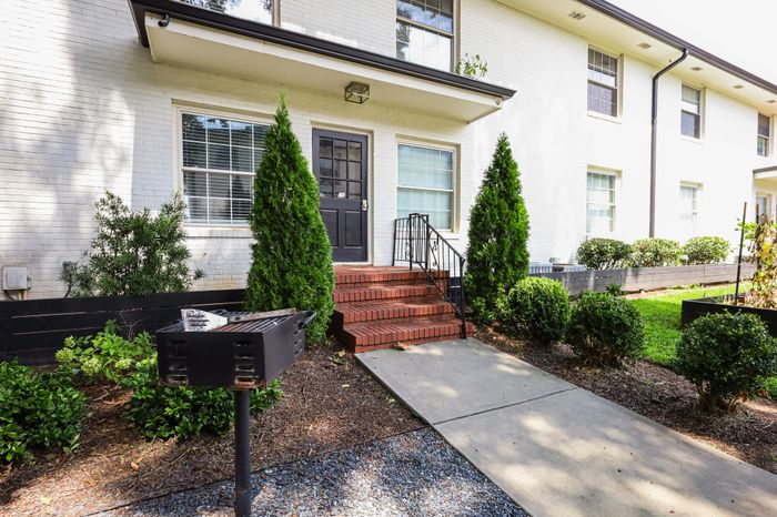 Charming apartment entrance with red brick steps, lush greenery, and a quaint grill in a serene setting.