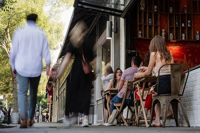 Vibrant city street scene with people enjoying outdoor dining at a trendy cafe, surrounded by lush trees and urban architecture.