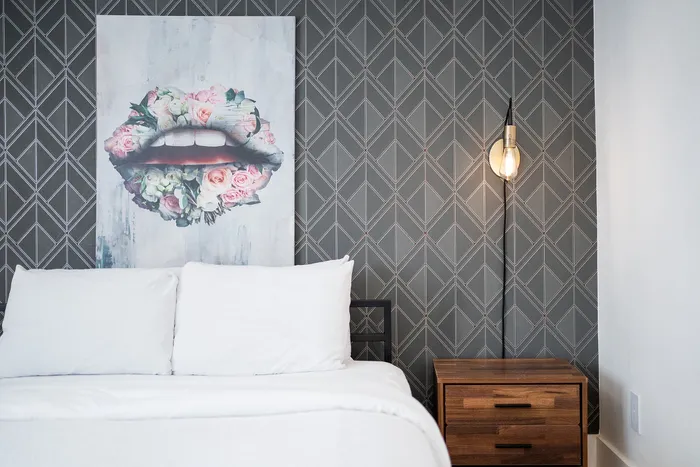 Chic bedroom featuring a modern geometric wallpaper, floral lips artwork, and cozy bedding alongside an elegant wooden nightstand with a stylish bulb lamp.