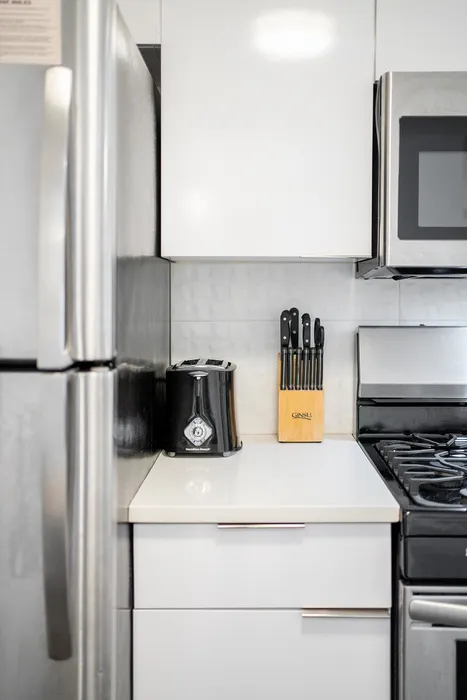 Sleek modern kitchen corner featuring a stainless steel refrigerator, knife block set, and contemporary black toaster, complemented by glossy white cabinets and a stylish stainless steel oven.