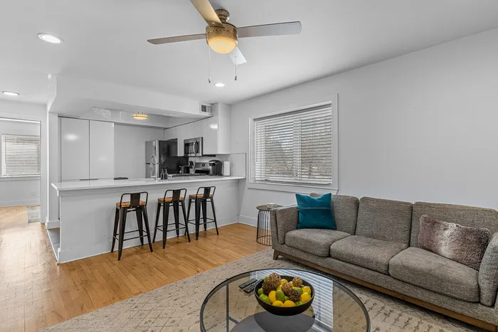 Modern open-plan living room and kitchen featuring sleek white cabinetry, a cozy gray sofa, and a stylish breakfast bar with chic stools. A ceiling fan adds comfort while a large window fills the space with natural light.