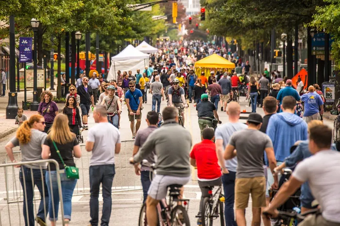 Vibrant urban scene with a bustling crowd enjoying a lively street festival, featuring cyclists and pedestrians against a backdrop of trees and tents.