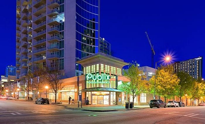Vibrant urban scene featuring a modern high-rise with a brightly lit Publix store at the base, surrounded by illuminated trees and bustling city life at dusk.
