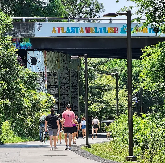 Vibrant urban pathway with people walking and cycling under a colorful Atlanta Beltline bridge, surrounded by lush greenery and street art.