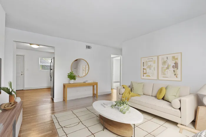 Bright and modern living room with elegant decor, featuring a comfortable cream sofa, chic coffee table, and stylish artwork, all set on polished hardwood floors. A welcoming entryway with a round mirror and neutral color palette enhances the open space.