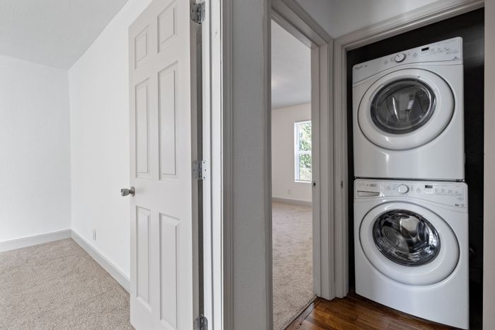Bright apartment hallway showcasing a modern stacked washer and dryer setup, leading into a cozy carpeted bedroom with natural light.