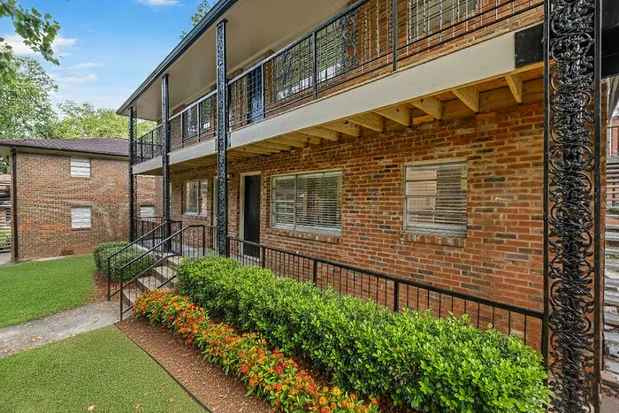 Charming brick apartment building with elegant wrought-iron details, lush landscaping, and expansive green lawn, offering serene and stylish living.