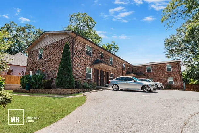 Charming brick apartment building with contemporary appeal, nestled in lush greenery, featuring ample parking space under a clear blue sky.