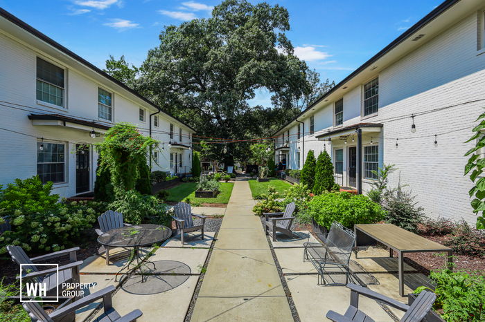 Charming courtyard view featuring elegant patio furniture, lush greenery, and white brick apartment buildings under a clear blue sky.