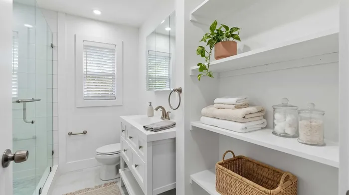 Bright, modern bathroom featuring a sleek walk-in shower, elegant vanity, and stylish open shelving with cozy towels and decorative jars.