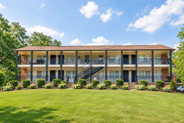 Charming two-story apartment building with spacious balconies, surrounded by lush greenery and vibrant landscaping under a bright blue sky.