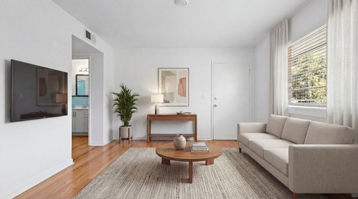 Bright and inviting living room with a modern beige sofa, sleek wooden coffee table, and a large wall-mounted TV. Natural light flows through sheer curtains, highlighting stylish decor and warm wood flooring.