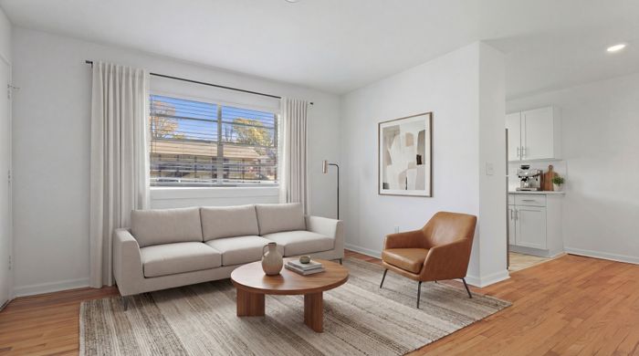 Bright and inviting living room with a modern beige sofa, chic wooden coffee table, and stylish brown armchair. Sunlight streams through large windows, complementing the elegant decor and open kitchen view.