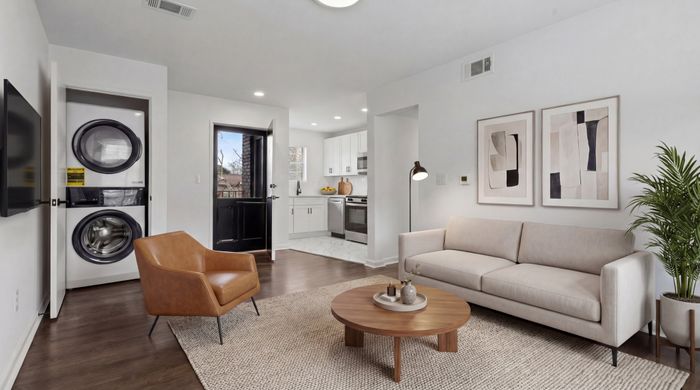 Contemporary living space with a cozy sofa, elegant armchair, and modern kitchen. Features include in-unit laundry and stylish wall art, offering a bright and inviting atmosphere.