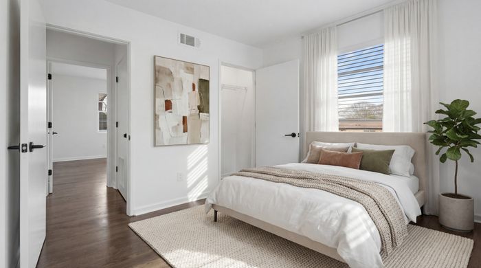 Bright and modern bedroom with stylish decor, featuring a cozy bed with plush pillows, elegant artwork, and sunlit windows with sheer curtains.