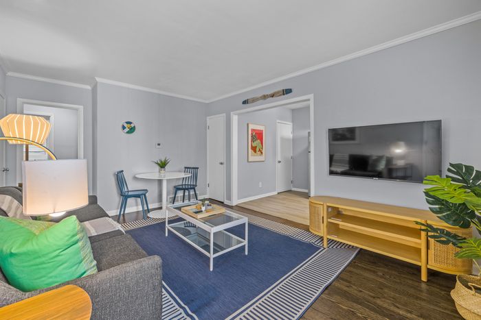 Bright and inviting living room with modern decor, featuring a cozy grey sofa, vibrant green cushions, stylish round dining table with blue chairs, and a sleek wall-mounted TV.