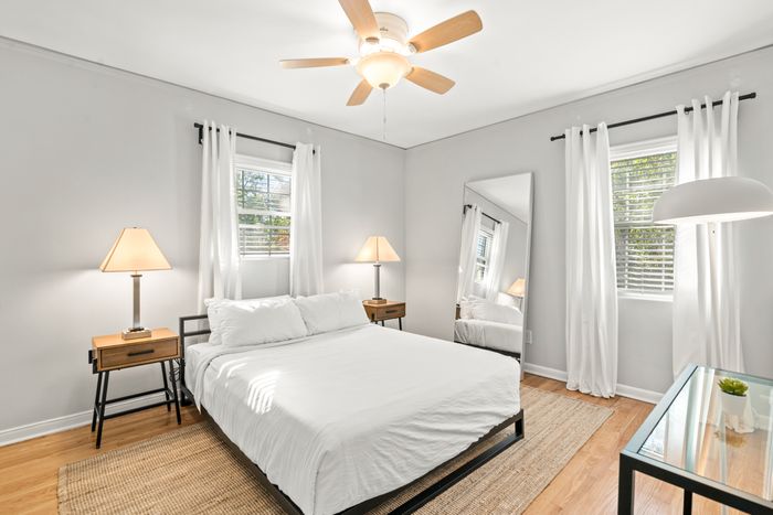 Bright and cozy bedroom featuring a comfortable bed, modern side tables with lamps, and a large wall mirror. The room is accentuated by natural light streaming through white-curtained windows onto polished hardwood floors.