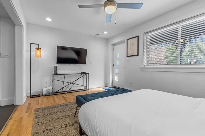 Inviting modern bedroom with sleek hardwood floors, featuring a wall-mounted flat-screen TV, stylish lamp, and large window for abundant natural light.
