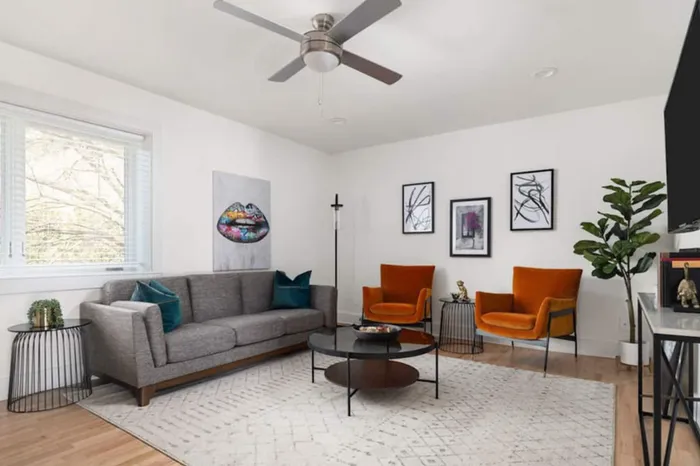 Chic and modern living room featuring a gray sofa, vibrant orange armchairs, stylish artwork, and lush greenery, all under a sleek ceiling fan.