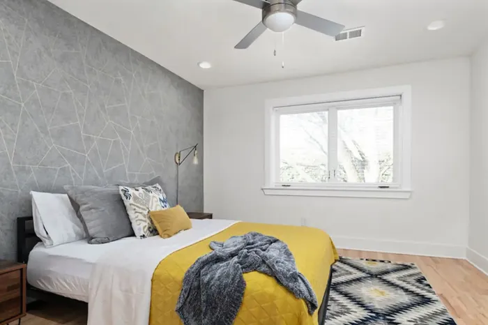 Bright and modern bedroom featuring a stylish gray accent wall, cozy bed with yellow and gray accents, and a large window offering natural light.