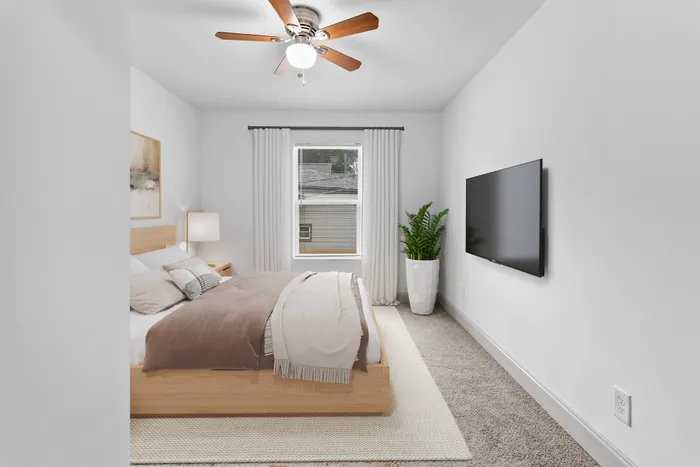 Bright and elegant bedroom with a cozy bed, modern ceiling fan, large window, and wall-mounted TV, creating a serene retreat.