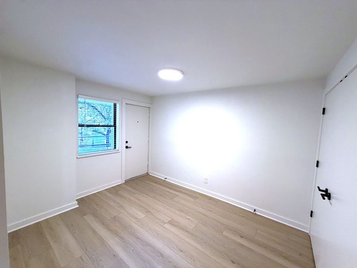 Bright and airy apartment room featuring sleek wooden flooring, a large window with natural light, and a modern entrance door.