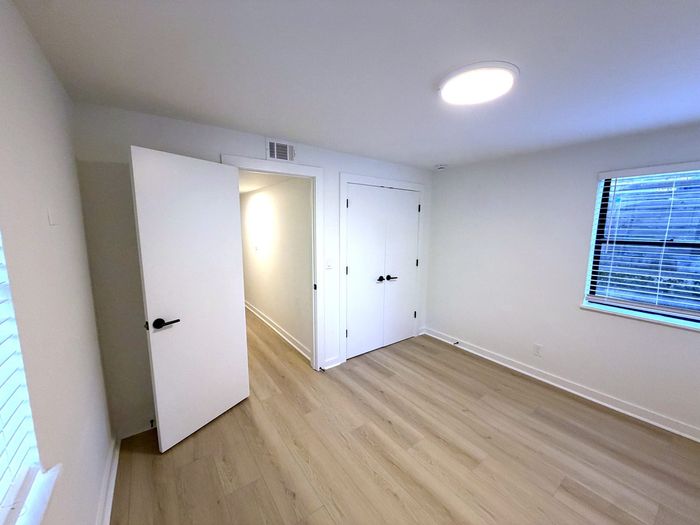 Bright and modern bedroom with light wood flooring, white walls, and dual doors leading to a hallway. Large window allows natural light to fill the space, creating a welcoming atmosphere.