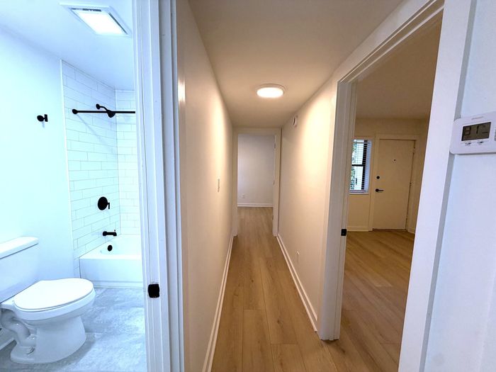Bright apartment hallway with sleek hardwood floors, leading to a modern bathroom with elegant tile work and stylish fixtures.