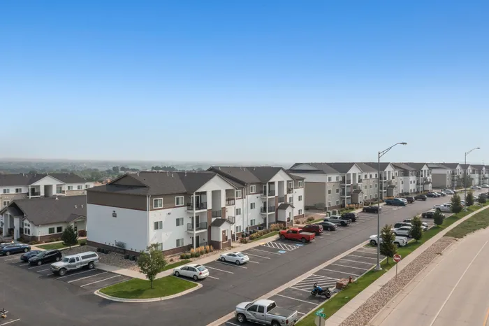Panoramic view of modern apartment complex with well-maintained parking and lush greenery against a clear blue sky backdrop.