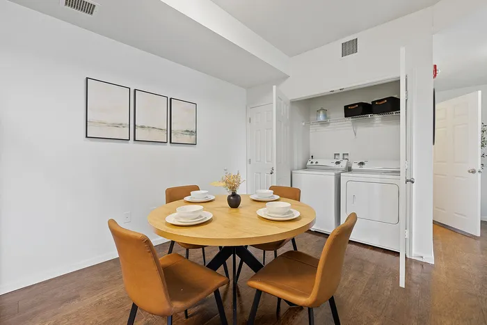 Chic dining area with contemporary round table set for four, adjacent to a well-organized laundry space with washer and dryer, accented by modern artwork and warm-toned wood flooring.