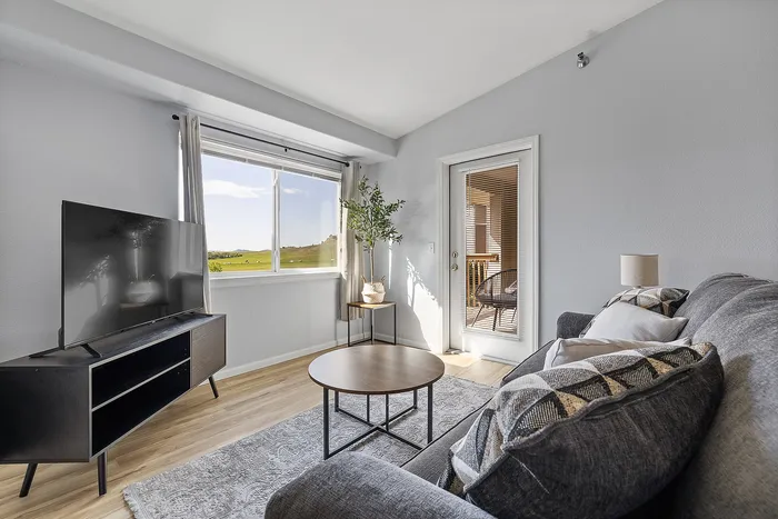 Bright and inviting living room with modern decor, featuring a comfortable sofa, elegant coffee table, and a flat-screen TV, complemented by a large window view of scenic greenery and a cozy balcony access.