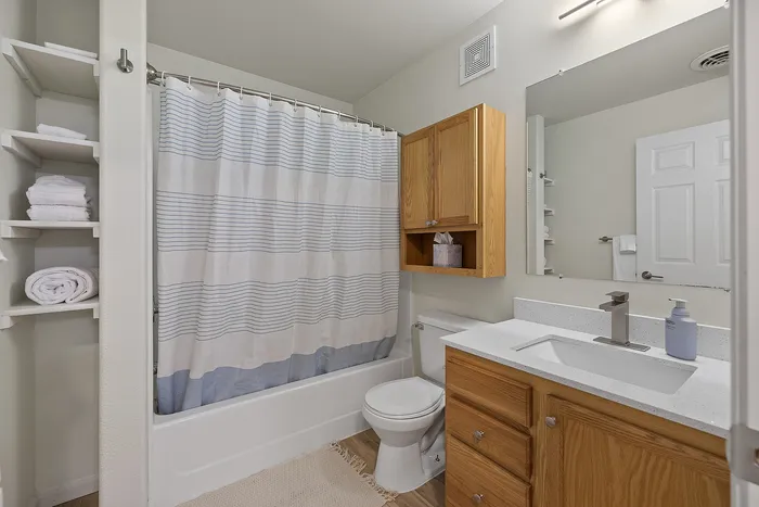 Elegant bathroom with modern fixtures, featuring a sleek white vanity, wooden cabinets, spacious mirror, and a refreshing shower with a striped curtain.