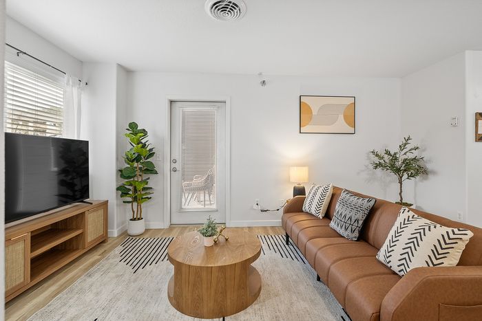 Contemporary living room with a chic brown leather sofa, stylish patterned cushions, and a modern wooden coffee table. Sunlit ambiance enhanced by large windows and indoor plants.