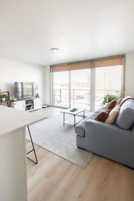 Bright living room with a modern gray sofa, large TV, and abundant natural light from floor-to-ceiling windows opening to a spacious balcony.