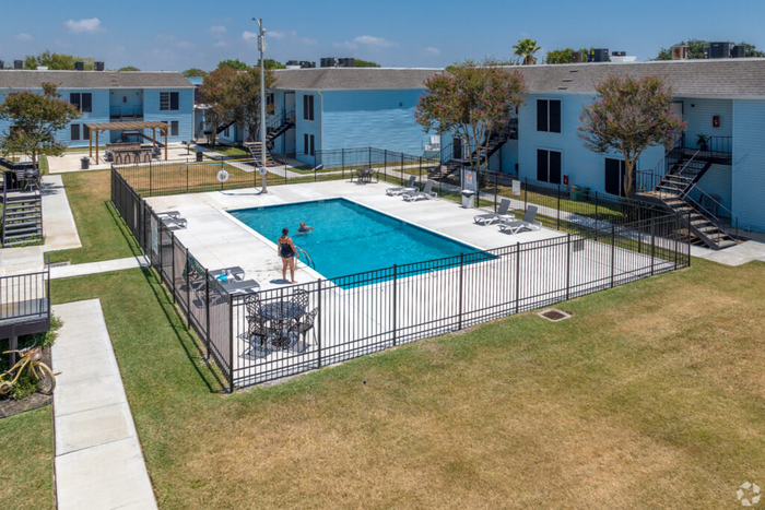 Inviting apartment community with a sparkling swimming pool, surrounded by well-maintained lawns and modern blue-toned buildings.