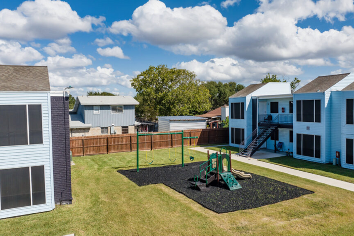 Charming apartment complex with serene landscaping and a vibrant children's play area under a bright blue sky.