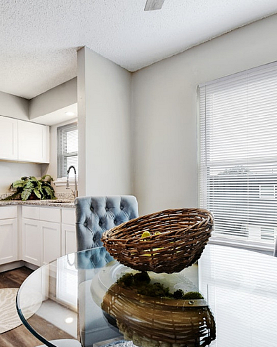 Bright and inviting kitchen with a glass-topped dining table, elegant tufted chair, and a decorative basket, illuminated by natural light through a large window.