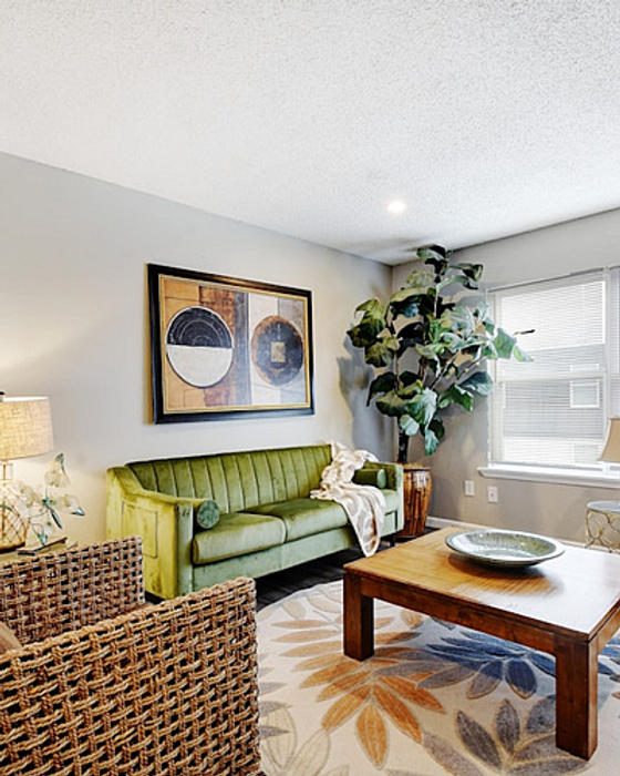 Bright and inviting living room with a stylish green sofa, modern art, lush plant, and a cozy wicker chair, ideal for relaxation and entertaining.