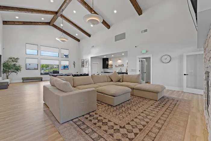 Spacious and elegant living room with high ceilings, exposed wooden beams, and large windows. Cozy beige sectional sofa on a patterned rug, adjacent to a modern kitchen.